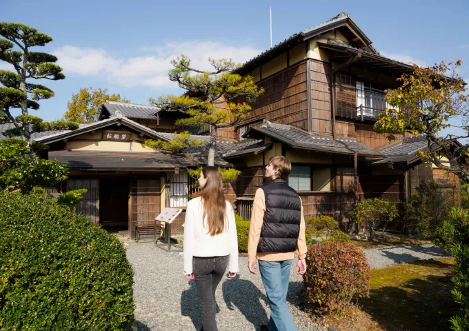 2人の男女が、砂利道と豊かな植栽のある伝統的な日本の木造家屋に入っていきます。