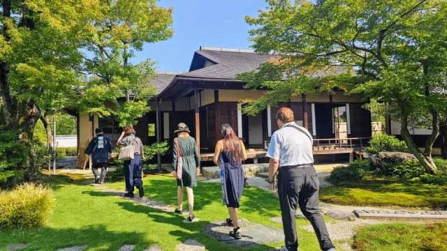 一群の人々が、伝統的な日本庭園を通り抜け、黒い瓦屋根の木造建築物に向かって歩いています。