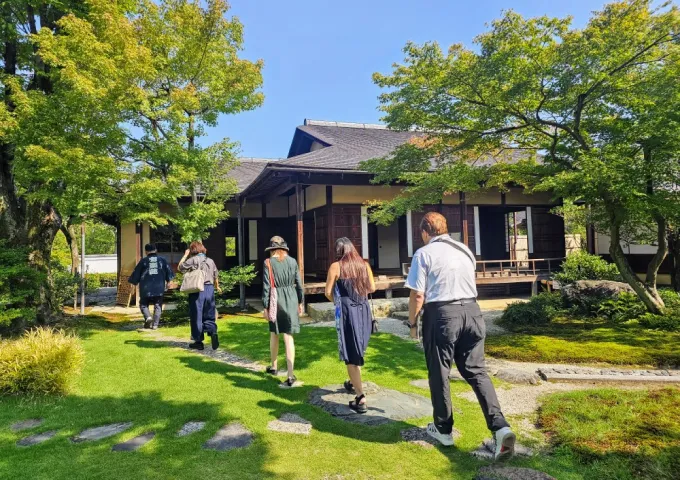 一群の人々が、伝統的な日本庭園を通り抜け、黒い瓦屋根の木造建築物に向かって歩いています。